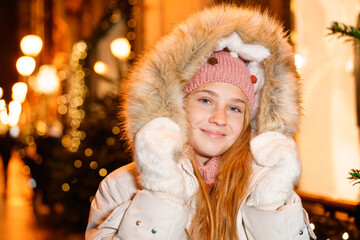 Street portrait smiling beautiful girl on festive christmas street in evening against background of christmas lights hands in mittens. Girl in stylish winter warm clothes. The model looks into camera.