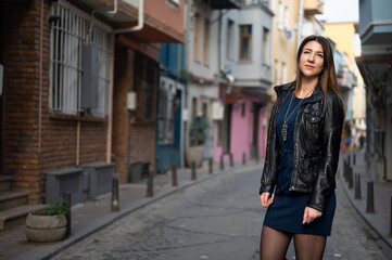 Tall attractive woman walks along a narrow city street. Young woman is wearing a leather jacket and short dress. look into the distance