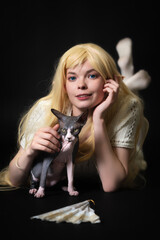 Young female elf cosplayer in white knitted dress, with long blonde hair lying down on stomach on black background, smiling and looking at camera. Sphynx kitten sits in front of her. Part of Series.