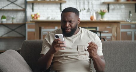 Surprised african man winner hold smartphone read good news amazed by mobile online bet bid game win at home. Happy male look at cell phone screen overjoyed by victory success while sitting on sofa.