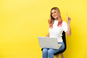 Young woman sitting on a chair with laptop over isolated yellow background showing and lifting a finger in sign of the best