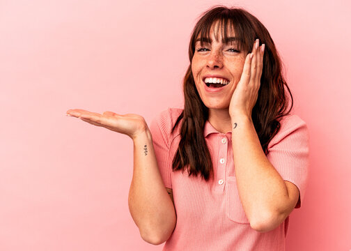 Young Argentinian woman isolated on pink background holds copy space on a palm, keep hand over cheek. Amazed and delighted.