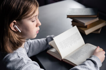 Serious little girl in headphones reads a book, blurred background.