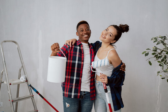 Handsome dark-skinned boy holds paint can and embraces his beautiful smiling woman having wall roller in hand, couple hugs, they renovate new apartment together, painting, finishing