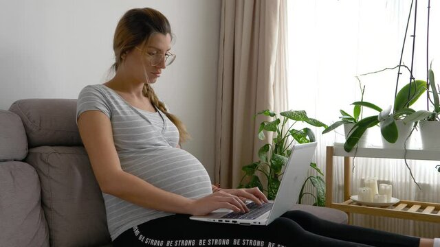 attractive pregnant woman types on laptop at home office couch