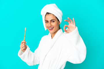 Young woman in a bathrobe brushing teeth over isolated blue background showing ok sign with fingers