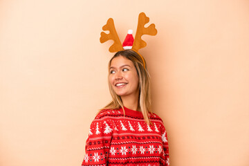 Young caucasian woman wearing a christmas reindeer hat isolated on beige background looks aside smiling, cheerful and pleasant.