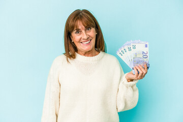 Middle age caucasian woman holding bank notes isolated on blue background happy, smiling and cheerful.