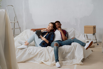 Smiling couple renting new apartment, renovating room, relaxing on sofa secured with foil attractive brunette lies on the sofa leaning against a dark-skinned boy holding a tablet in her hands