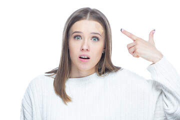 Close up shot of young scared woman pointing with her finger on an applied adhesive bandage on her forehead after injury isolated on white background