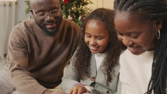 Tilt up close up of happy African-American father, mother and 10-year-old daughter sitting on floor in living room on Boxing Day and petting adorable maltipoo dog. Christmas tree in background