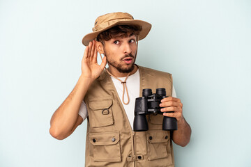 Young mixed race man holding binoculars isolated on blue background trying to listening a gossip.