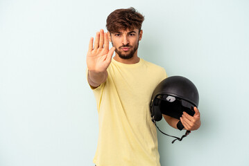 Young mixed race man holding motorcycle helmet isolated on blue background standing with outstretched hand showing stop sign, preventing you.