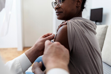 Shot of healthcare worker injecting black female patient with COVID-19 vaccine. Beautiful young black girl takes seasonal flu jab. Black woman getting injection at home. Immunization, COVID-19 vaccine - obrazy, fototapety, plakaty