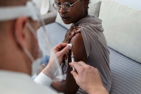 Woman receiving coronavirus vaccine. Shot of an African American woman getting vaccinated by a doctor at home. Young doctor man injecting COVID-19 vaccine for woman sitting on the sofa at clinic
