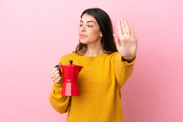 Young Italian woman holding a coffee maker isolated on pink background making stop gesture and disappointed