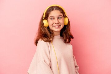 Little caucasian girl listening to music isolated on pink background looks aside smiling, cheerful and pleasant.