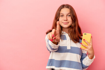 Little caucasian girl holding mobile phone isolated on pink background showing number one with finger.