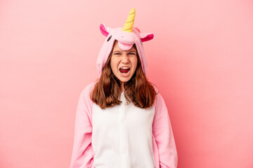 Little caucasian girl wearing a unicorn pajama isolated on pink background screaming very angry and aggressive. © Asier