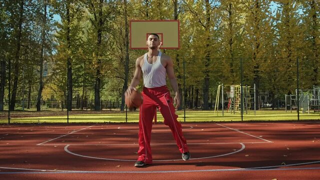 Young sportsman posing with basketball ball in sunlight. Portrait of handsome basketball player outdoors. Mixed race man looking at camera. Full length shot in 4K, UHD