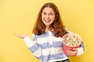 Little caucasian girl holding popcorns isolated on yellow background showing a copy space on a palm and holding another hand on waist.