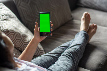 A young woman sitting on the couch is using a smartphone with a pre-configured green screen.