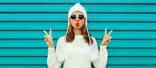 Winter portrait of beautiful young woman blowing her lips sending air kiss wearing a white knitted sweater, hat on blue background