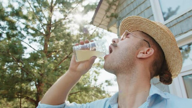 Old fashioned fancy young hipster man with moustache in straw hat, pink sunglasses drink fresh pineapple juicein villgae summer country house. Morining of modern outdoor freelance lifestyle
