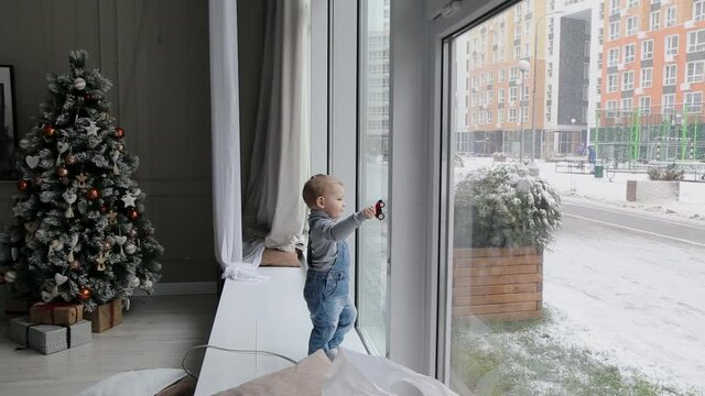 Little Boy plays by the window in the winter in a car in an apartment with a beautiful decorated Christmas tree on Christmas morning.
