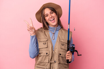 Young caucasian fisherwoman holding a rod isolated on pink background joyful and carefree showing a peace symbol with fingers.
