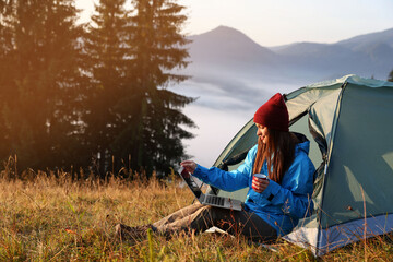 Woman working on laptop near camping tent outdoors surrounded by beautiful nature - obrazy, fototapety, plakaty