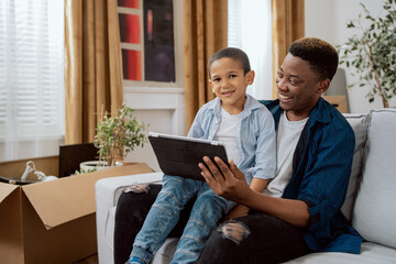 A smiling man holds a tablet in his hand, shows his son videos, funny pictures, they browse the Internet, boys laugh, father and son spend time together on the couch