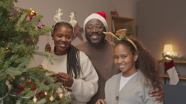 Portrait of happy African-American family of three wearing festive hairbands and Santa hat posing for camera while decorating Christmas tree at home