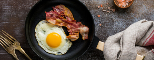Traditional English breakfast with fried eggs and bacon in cast iron pan on dark concrete background. Top view.