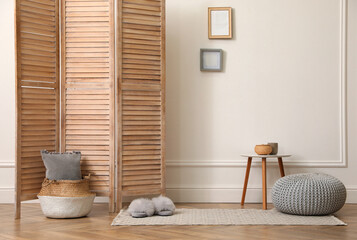Folding screen, small table and knitted pouf near white wall in room. Interior design
