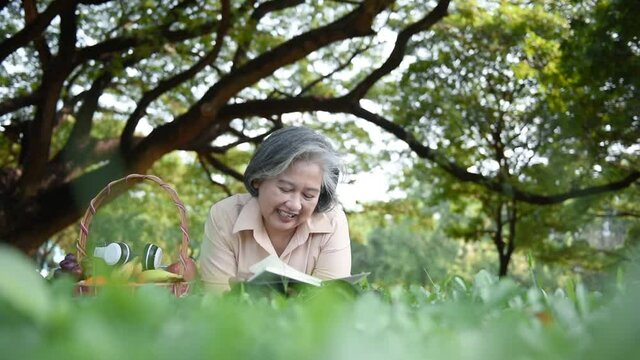 Asian elderly woman, lying and relaxing on the green lawn in garden and reading a notebook diary Which recorded her story with happy, to retirement age and nature concept.