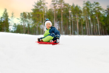 Fototapeta na wymiar childhood, sledging and season concept - happy little boy sliding on sled down snow hill outdoors in winter over snowy park or forest background