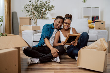 Obraz premium Married couple after moving to new house unpack boxes sitting on floor, woman shows husband found photo in frame from their wedding, man embraces wife, they smile reminisce