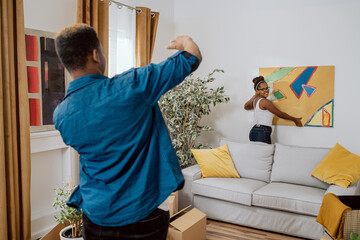 A woman hangs a painting on wall in new apartment, together with her boyfriend they assess if it...