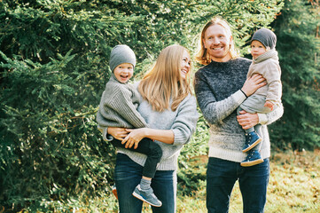 Outdoor portrait of beautiful happy young family posing in pine forest, wearing warm pullovers,...