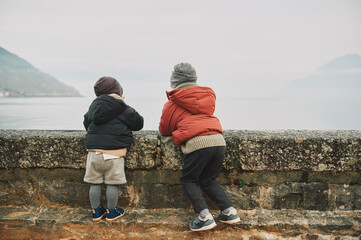 Outdoor portrait of two funny kids playing by the lake on a cold autumn day, wearing warm winter...