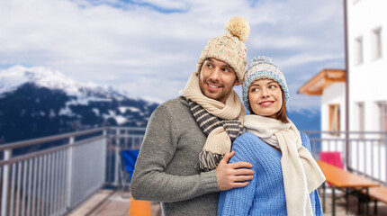 travel, tourism and winter holidays concept - happy couple in knitted hats and scarves over mountains and ski resort background
