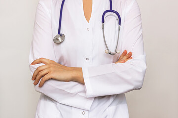 Cropped shot of a young female doctor in a white coat with a stethoscope around her neck stands with her hands crossed on her stomach isolated on a gray background