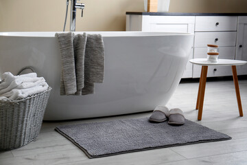 Soft grey mat with slippers on floor near tub in bathroom. Interior design
