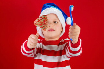 Happy Christmas. Blonde boy kid in a Santa's hat with a candy and a blue toothbrush in his hands on red background. To choose. Place for text. Dental hygiene, children's health concept.