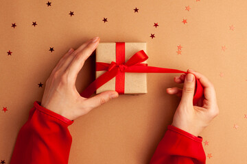 Human hands with red sleeves untiding a brown paper gifts with a red satin ribbon bow on brown background with red stars shapes. Christmas Holidays concept flat lay.