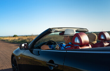 Elderly man and woman in straw hats in black cabriolet with red salon with open roof in summer on country road with clear blue sky