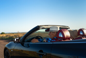 Fototapeta na wymiar Elderly man and woman in straw hats in black cabriolet with red salon with open roof in summer on country road with clear blue sky