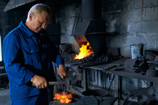 Side view of senior man in special dark blue uniform working with special tool and creating metal near heating equipments. Concept of process creating fence in smithy.