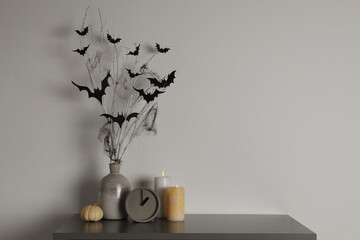 Alarm clock, burning candles and vase with paper bats  on table near white wall, space for text. Halloween decor